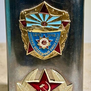 May include: A silver-colored flask with a textured cap. The flask features two ornate emblems: a star with a hammer and sickle, and a badge with text and a starburst design. The emblems are red, gold, and blue.