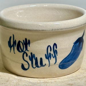 May include: A small, off-white ceramic bowl with a blue hand-painted eggplant and the words "Hot Stuff" in blue script. The bowl has a rounded shape and a slightly flared rim, suitable for serving dips or sauces.