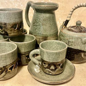May include: A collection of celadon green ceramic teaware with a crackle glaze. The set includes a teapot with a woven handle, a pitcher, cups, and saucers. The cups have decorative cutouts and a dark brown band.