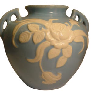 Könnte beinhalten: Eine hellblaue Keramikvase mit weißen Blumenmustern. Die Vase hat zwei Henkel und einen runden Körper.