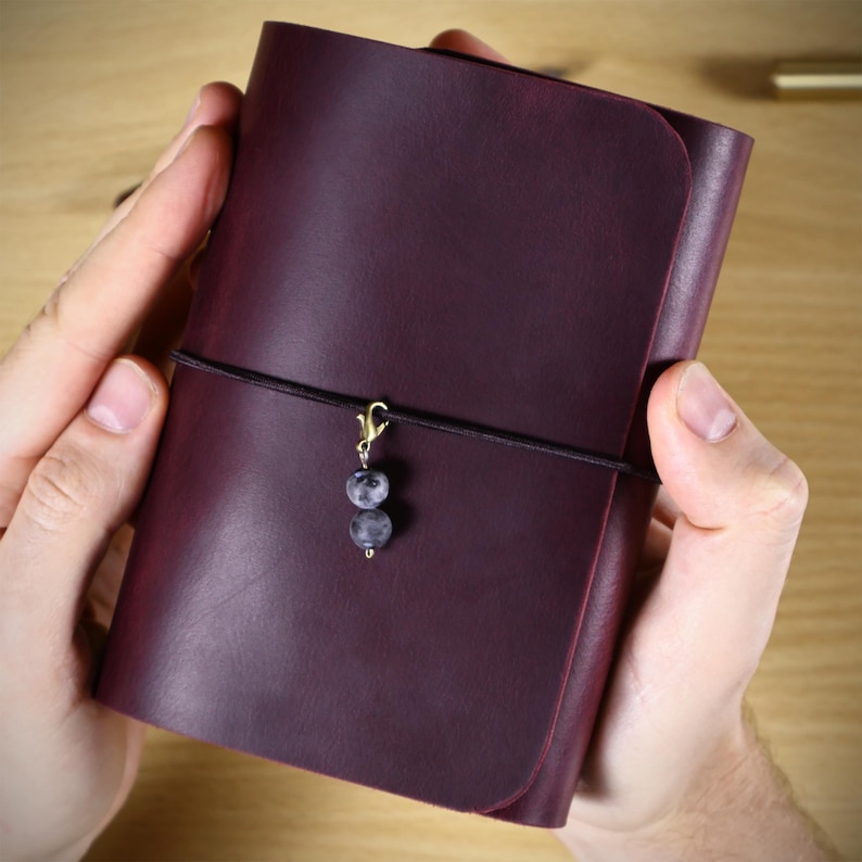 May include: A deep purple leather journal cover featuring a smooth, matte finish. A thin black elastic cord wraps horizontally around the center, secured with a gold-toned lobster clasp holding two small, round, mottled grey stone beads.