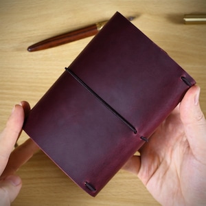 May include: A deep burgundy leather journal cover featuring a smooth, matte finish and a thin black elastic cord wrapped horizontally around the center to secure the cover. The rectangular journal has rounded corners and visible stitching details.