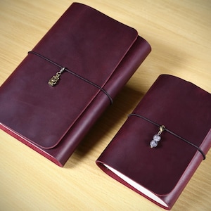 May include: Two deep burgundy leather journals with wrap-around black elastic cords. The larger journal features a small brass-toned lantern charm, while the smaller journal includes a charm with two small, light-colored spherical beads.