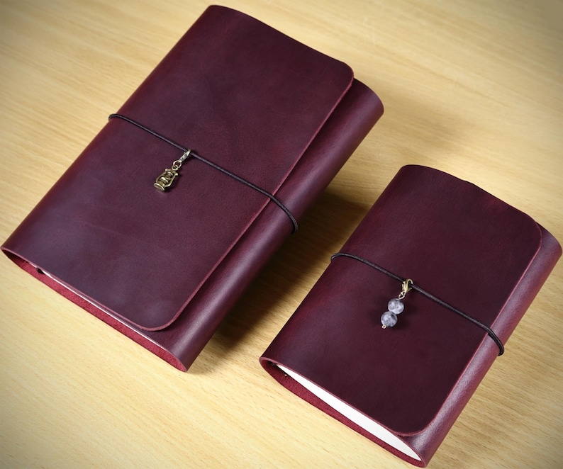 May include: Two deep burgundy leather journals with wrap-around black elastic cords. The larger journal features a small brass-toned lantern charm, while the smaller journal includes a charm with two small, light-colored spherical beads.