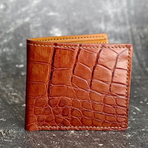May include: A brown bi-fold wallet with a textured, crocodile-like pattern. The wallet is open, revealing a lighter brown interior. The edges are stitched with a contrasting thread. A classic accessory.
