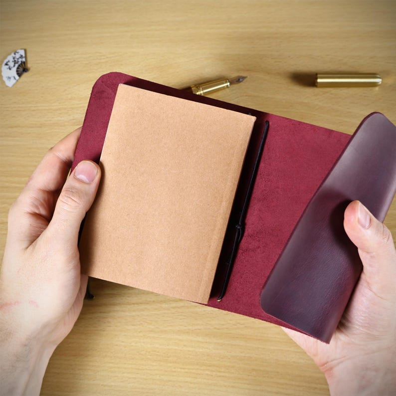 May include: A deep burgundy leather journal cover featuring a smooth exterior and a soft, suede-like interior. The cover holds a rectangular notebook with light brown, unlined paper pages secured by a thin black elastic cord running vertically.