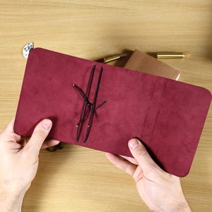 May include: A deep burgundy suede leather journal cover featuring a rectangular shape with rounded corners and a central black elastic cord system designed to hold inserts in place. The cover has a soft, matte texture and a minimalist design.