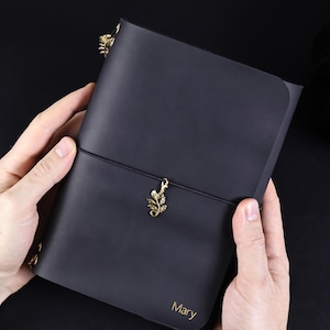 Könnte beinhalten: Ein mattschwarzes Notizbuch mit glattem Einband, das unten rechts den in Gold geprägten Namen Mary trägt. Ein dünnes schwarzes Gummiband umschließt die Mitte und hält in der Mitte einen kleinen, goldfarbenen Metallanhänger in Blattform.