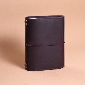 May include: A dark brown leather journal cover with a smooth matte finish. The cover features a thin, dark red elastic band wrapped horizontally around the center to keep it closed, and gold-toned metal eyelets at the spine for binding.