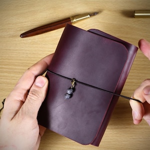 May include: A deep purple leather journal with a wrap-around black elastic cord closure. The cord features a small metal clasp adorned with two round, mottled grey stone beads. The journal has a smooth, matte finish and rounded corners.