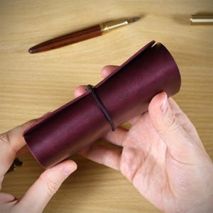 May include: A cylindrical dark burgundy leather pen case secured with a thin black elastic cord. The smooth, matte-finish leather is wrapped into a roll shape, providing a protective sleeve for writing instruments.