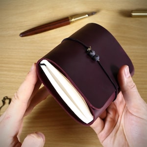 May include: A dark purple leather journal cover featuring a black elastic closure band with two small grey spherical beads. The cover wraps around a stack of white paper pages, creating a compact and secure notebook design for writing or sketching.