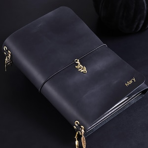 Könnte beinhalten: Ein schwarzes Lederjournal mit dem in Gold geprägten Namen Mary auf dem Vorderdeckel. Das Journal wird durch ein schwarzes Gummiband mit einem goldenen Blattanhänger verschlossen, weitere Blattanhänger sind an der seitlichen Bindung befestigt.