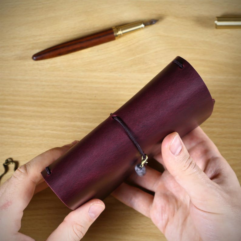 May include: A deep purple leather journal cover with a smooth texture, secured by a thin black elastic cord featuring a small, round, grey stone charm. The cover is cylindrical in shape and is held in place by the elastic band wrapped around it.
