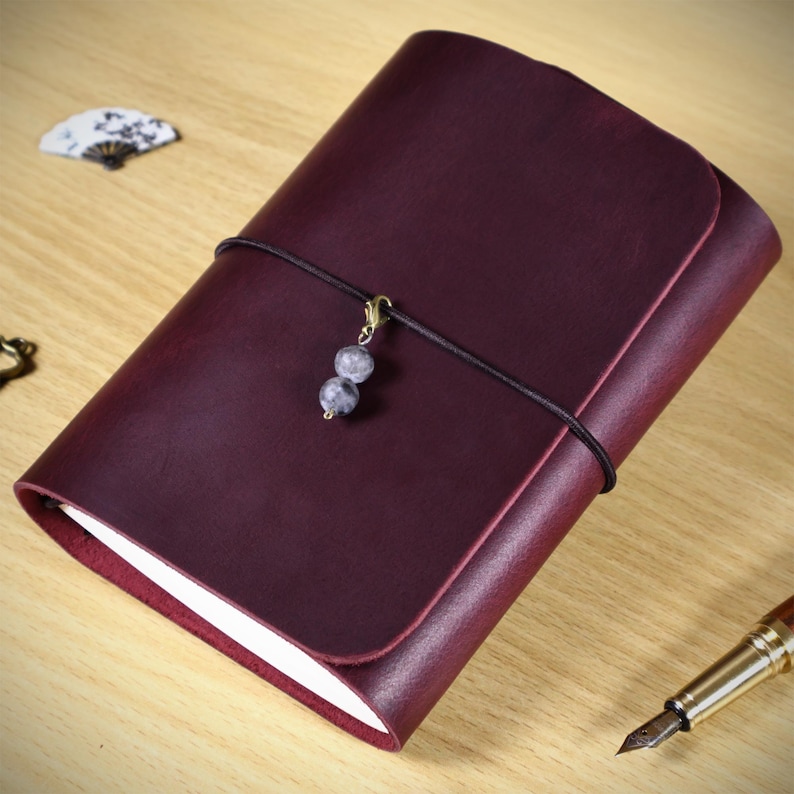 May include: A deep burgundy leather journal with a smooth matte finish, secured by a thin black elastic cord. A small gold-toned clasp hangs from the cord, featuring two round, mottled grey and white stone beads as a decorative accent.