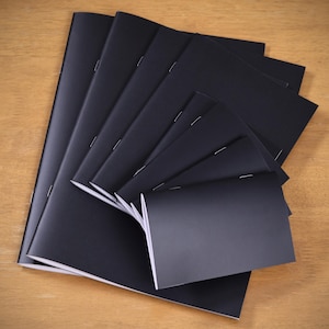 Op de afbeelding: Een verzameling zwarte notitieboekjes, uitgespreid op een houten oppervlak. De notitieboekjes hebben een eenvoudig, minimalistisch ontwerp met een matte afwerking. De pagina's zijn wit en de binding is met nietjes.
