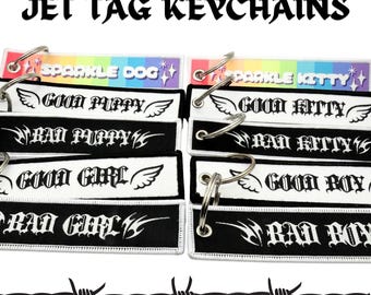 May include: A collection of rectangular embroidered jet tag keychains featuring black and white fabric with gothic-style text including Good Puppy, Bad Puppy, Good Kitty, Bad Kitty, Good Girl, Bad Girl, Good Boy, and Bad Boy, plus rainbow Sparkle Dog and Sparkle Kitty tags.