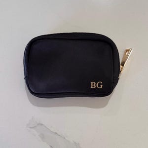 Peut inclure: Petite pochette bleu marine avec une fermeture éclair dorée et les initiales "BG" en lettres dorées. La pochette est rectangulaire avec des bords arrondis, et la tirette de la fermeture éclair est également dorée. Elle est pour cosmétiques ou accessoires.