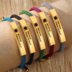 Puede incluir: Seis pulseras de alerta médica con placas rectangulares doradas y cordones coloridos. Cada placa tiene el texto "Mark Doratti BCMed# 9767361277" y un símbolo médico. Los cordones son azules, negros, verdes, magenta, rojos y grises.