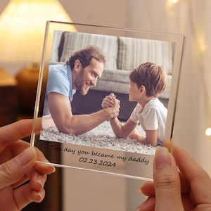May include: A clear glass photo frame with a picture of a father and son arm wrestling. The frame has the text "The day you became my daddy 02.23.2024" inscribed on the bottom.