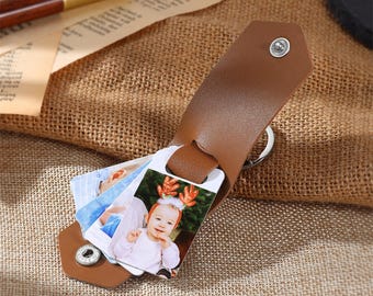 May include: Brown leather photo keychain with a metal ring and a snap closure. The keychain holds multiple photo cards, one of which features a baby wearing a reindeer headband.