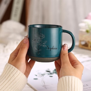 May include: A teal blue ceramic mug with a floral design and the name "Sophia" engraved on the side. The mug has a brown base.