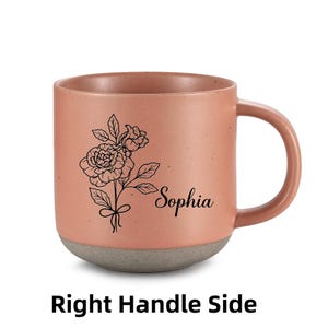 May include: A pink ceramic mug with a gray base. The mug has a black line drawing of a flower and the name "Sophia" on the front. The mug has a handle on the right side.