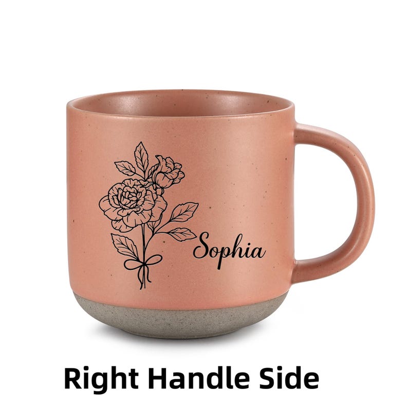 May include: A pink ceramic mug with a gray base. The mug has a black line drawing of a flower and the name "Sophia" on the front. The mug has a handle on the right side.