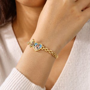 May include: A gold-toned metal link bracelet featuring a heart-shaped locket charm. The locket is partially open, revealing a small, rectangular photograph of a couple inside. The bracelet consists of small, repeating rectangular links.