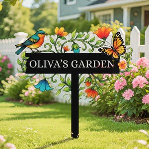 Puede incluir: Un letrero de jardín de metal negro con el texto "OLIVA'S GARDEN". El letrero presenta un pájaro colorido, una mariposa, flores y enredaderas. El letrero está sobre un poste negro y está colocado en un jardín.