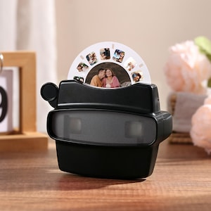 Puede incluir: Un visor estereoscópico de plástico negro con un disco fotográfico circular blanco insertado en la ranura superior. El dispositivo tiene dos lentes rectangulares en la parte frontal y una palanca negra redonda en el lado izquierdo para avanzar el disco.