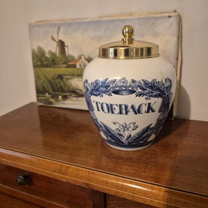 Peut inclure: Un pot en céramique blanc avec un couvercle doré et un motif floral bleu et blanc. Le mot "TOEBACK" est imprimé sur le devant. Le pot est posé sur une surface en bois.