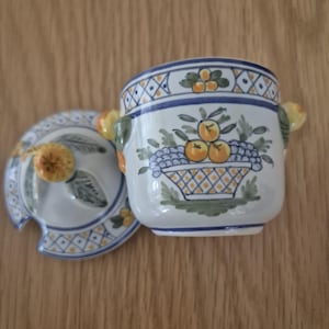 Puede incluir: Un azucarero de cerámica blanca con tapa, con un diseño floral azul y amarillo. El cuenco tiene una ilustración de una cesta de frutas y pequeñas asas amarillas. La tapa tiene un diseño a juego y un asa con forma de fruta amarilla.