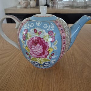 Op de afbeelding: Een keramische theepot met een lichtblauwe achtergrond en een groot roze rozenbloemmotief. De theepot heeft een wit handvat en tuit, en een wit deksel met een kleine knop. De theepot is versierd met andere bloemen- en kantpatronen.