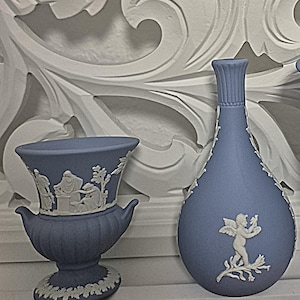 Może przedstawiać: Jasnoniebieski wazon ceramiczny z szerokim brzegiem i dwoma uchwytami, ozdobiony białymi figurami w reliefie przedstawiającymi ludzi, obok jasnoniebieskiej butelki ceramicznej z wąską szyjką i białą figurą w reliefie skrzydlatego cherubina z ptakiem.