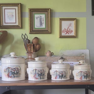 Pode incluir: Um conjunto de quatro potes de cerâmica na cor creme com tampas, cada um apresentando uma ilustração colorida de figuras dançando em uma vila. Os potes variam em tamanho, dispostos do maior para o menor, e incluem alças laterais em cada peça.