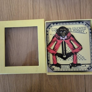 Könnte beinhalten: Ein Vintage-Multiplikationsspiel "Educated Monkey" in einer gelben Box. Das Spiel zeigt einen Cartoon-Affen in einem roten Mantel und einer Fliege mit Multiplikationstabellen. Die Box ist geöffnet und zeigt das Spielstück.