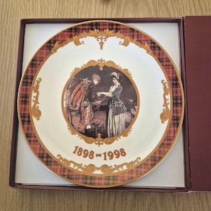 Puede incluir: Plato decorativo con una imagen central de una pareja con vestimenta de época, rodeado por un borde a cuadros rojo y negro. Detalles dorados y el texto "1898-1998" están presentes. El plato se presenta en una caja granate.