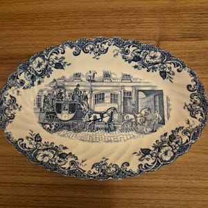Peut inclure: Plat ovale en céramique avec un motif bleu et blanc. L'image centrale représente une diligence tirée par des chevaux devant un bâtiment. La bordure présente des motifs floraux et des volutes. Environ 30 cm de long.