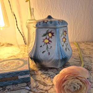 Könnte beinhalten: Ein Vintage-Keramiktopf mit Deckel, verziert mit roten, gelben und grünen Blumenmustern auf hellblauem und weißem Grund. Der Topf steht auf einer Marmoroberfläche, neben einer kleinen dekorativen Schachtel und einer rosa Blume.