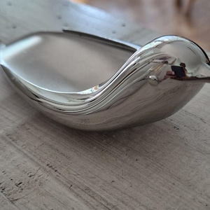 Könnte beinhalten: Eine polierte silberne Vogelschale. Die Schale hat eine glatte, reflektierende Oberfläche und ein geschwungenes Design mit einer schnabelartigen Form an einem Ende. Die Schale ist wahrscheinlich aus Metall und steht auf einer Holzoberfläche.