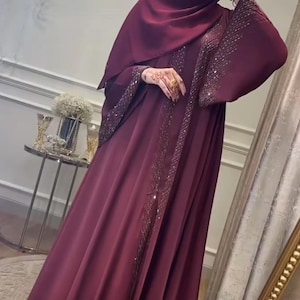 May include: A burgundy abaya with embellished sleeves and trim. The flowing garment features a matching head covering. The abaya is made of a smooth, draping fabric, and is suitable for modest wear.