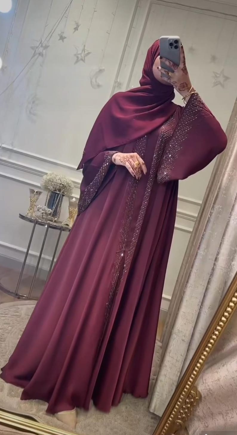 Op de afbeelding: Een diepbordeauxrode abaya met lange, vloeiende mouwen en een bijpassende hijab. Het kledingstuk is versierd met ingewikkeld, glinsterend kralenwerk en pailletten langs de opening aan de voorzijde en de mouwboorden, wat zorgt voor een textuur.