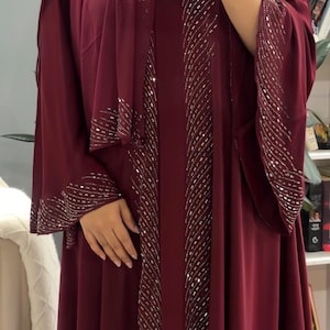 Op de afbeelding: Een diepbordeauxrode abaya met lange, wijde mouwen en een bijpassende hoofddoek. Het kledingstuk is versierd met verticale rijen zilverkleurige metalen versieringen en pailletten langs de vooropening en de manchetten van de mouwen.