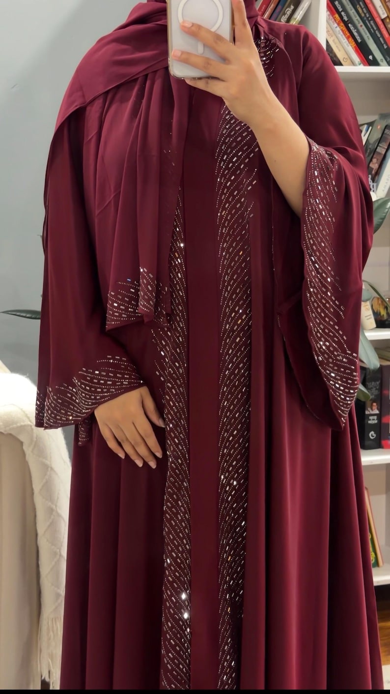 Op de afbeelding: Een diepbordeauxrode abaya met lange, wijde mouwen en een bijpassende hoofddoek. Het kledingstuk is versierd met verticale rijen zilverkleurige metalen versieringen en pailletten langs de vooropening en de manchetten van de mouwen.