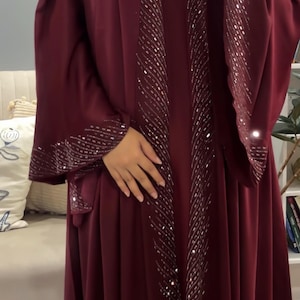 Op de afbeelding: Een diepbordeauxrode abaya met lange, wijde mouwen en een open voorzijde. Het kledingstuk is versierd met ingewikkelde zilveren pailletten en kralen die de randen van de opening aan de voorzijde en de manchetten van de mouwen omlijnen.