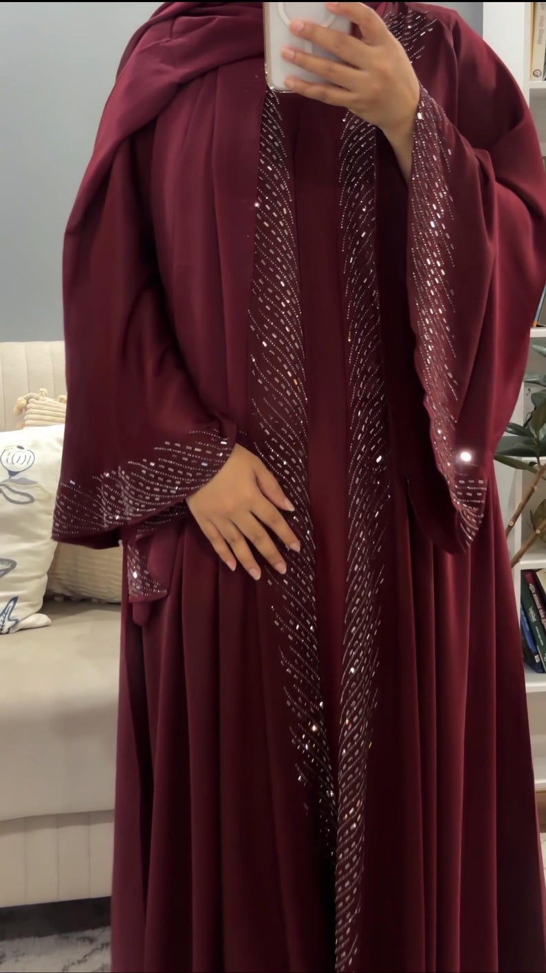 Op de afbeelding: Een diepbordeauxrode abaya met lange, wijde mouwen en een open voorzijde. Het kledingstuk is versierd met ingewikkelde zilveren pailletten en kralen die de randen van de opening aan de voorzijde en de manchetten van de mouwen omlijnen.