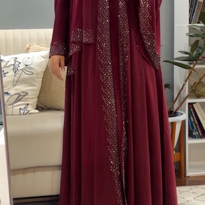 Op de afbeelding: Een bordeauxrode abaya die tot op de grond reikt, inclusief een bijpassende hijab en een open ontwerp aan de voorzijde. Het kledingstuk is versierd met verticale rijen zilveren lovertjes langs de voorranden en mouwboorden voor een glinsterend effect.