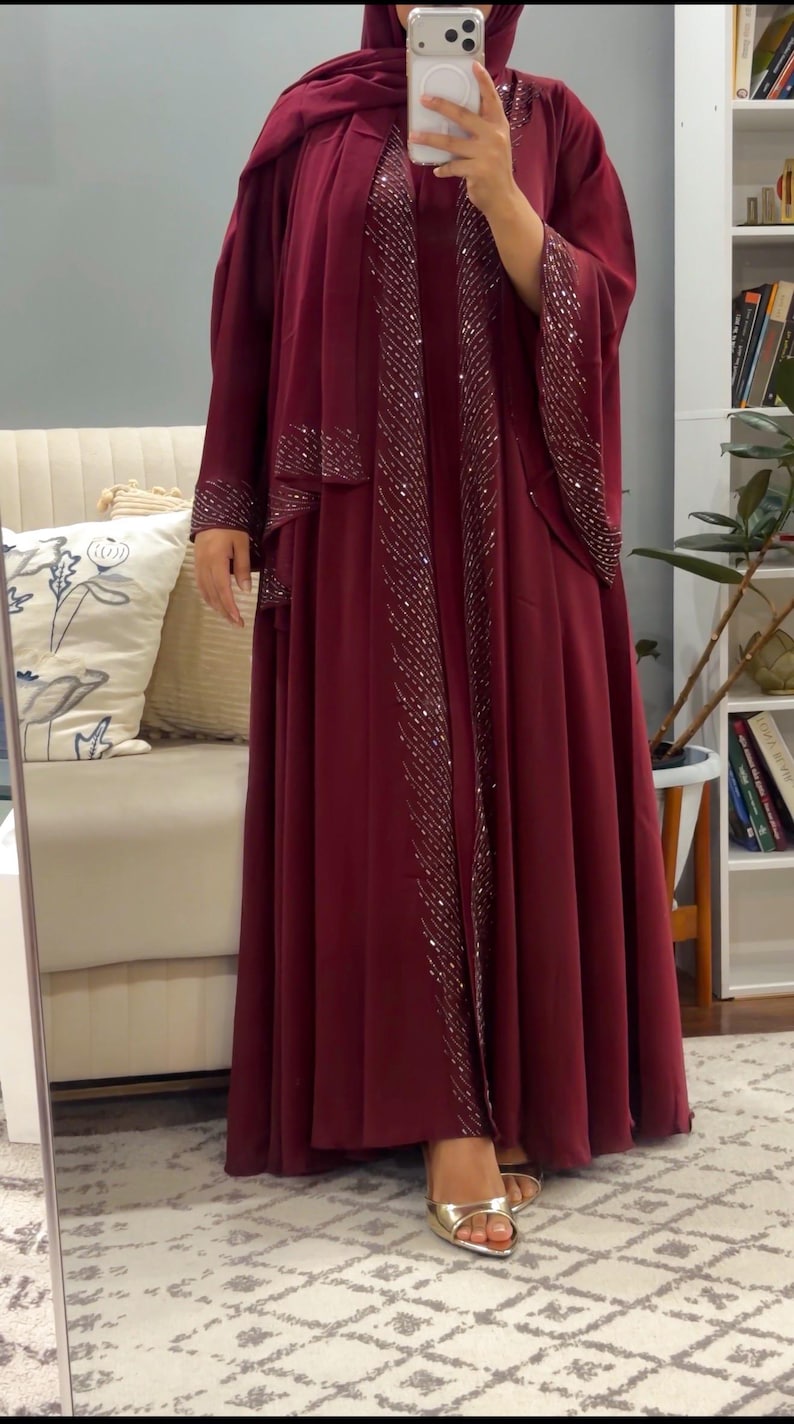 Op de afbeelding: Een bordeauxrode abaya die tot op de grond reikt, inclusief een bijpassende hijab en een open ontwerp aan de voorzijde. Het kledingstuk is versierd met verticale rijen zilveren lovertjes langs de voorranden en mouwboorden voor een glinsterend effect.