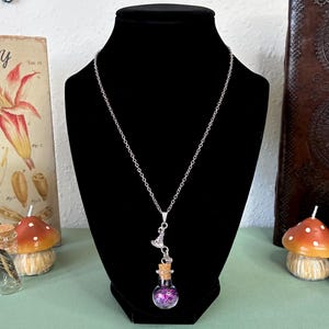 May include: A silver-toned chain necklace featuring a small glass vial pendant filled with purple and pink glitter. The pendant is sealed with a cork stopper and accented with a small silver-toned witch hat charm attached to the top of the vial.
